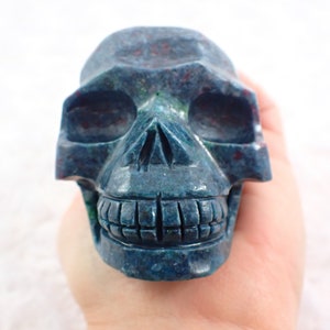 Peut inclure: Une figurine de crâne sculpté dans des tons de bleu et de vert avec des accents rouges. Le crâne présente des détails, notamment des orbites, une cavité nasale et une rangée de dents. La figurine est tenue dans une main.