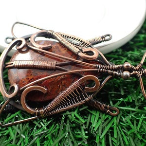 May include: A copper wire wrapped pendant featuring a red and brown agate stone. The pendant is shaped like a stylized insect with intricate wirework details.