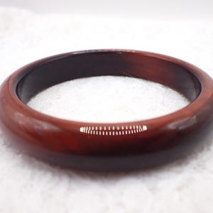 May include: A polished, round bangle bracelet in shades of brown. The bracelet has a smooth, glossy finish and a dark interior. The overall design is simple and elegant, suitable for everyday wear. The bracelet's diameter is approximately 2.75 inches.