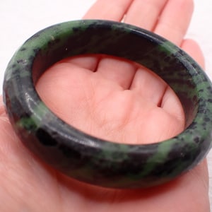 May include: A dark green and black jade bangle bracelet. The bracelet is a solid, circular band with a polished finish. The jade has a mottled pattern of dark green and black throughout.