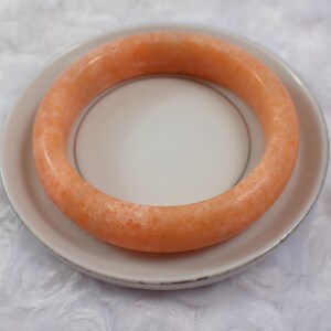 May include: An orange-colored bangle bracelet rests on a white plate with a silver rim. The bracelet has a smooth, polished surface and a solid, circular design. The plate is set against a white, textured background.