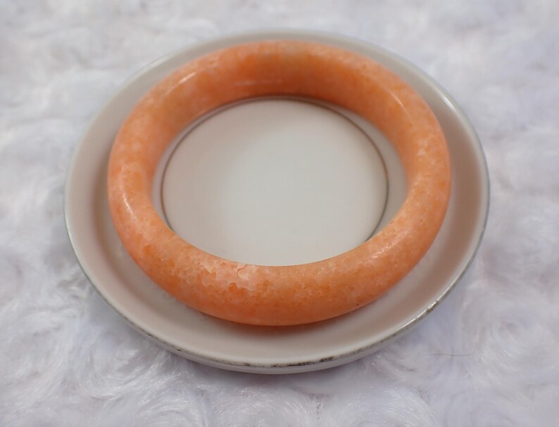 May include: An orange-colored bangle bracelet rests on a white plate with a silver rim. The bracelet has a smooth, polished surface and a solid, circular design. The plate is set against a white, textured background.