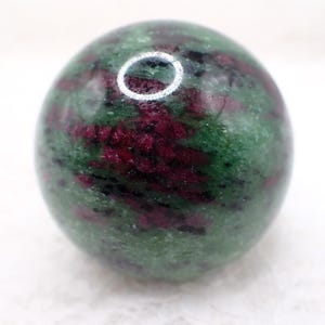 Könnte beinhalten: Eine polierte Kugel aus Rubin in Zoisit. Der Edelstein weist ein gesprenkeltes Muster aus tiefroten und grünen Farbtönen auf. Die Kugel ist glatt und rund, mit einem glänzenden Finish. Die Farben sind gut vermischt und erzeugen ein einzigartiges und natürliches Aussehen.