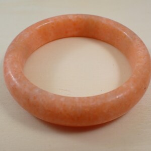 May include: A peach-colored bangle bracelet. The round bracelet is made of a solid, opaque material with a slightly speckled appearance. The bracelet is a simple, elegant piece of jewelry.