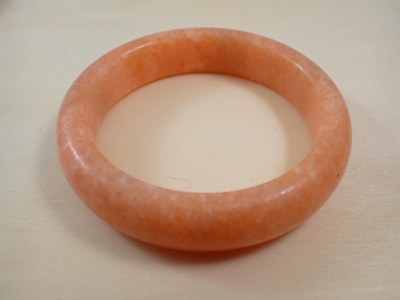 May include: A peach-colored bangle bracelet. The round bracelet is made of a solid, opaque material with a slightly speckled appearance. The bracelet is a simple, elegant piece of jewelry.
