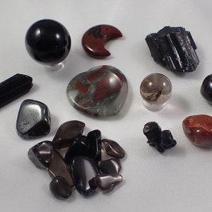 May include: A collection of black, red, and clear crystals and stones, including a black obsidian sphere, a red jasper heart, a black tourmaline point, and a clear quartz sphere.