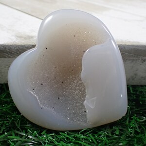 May include: A heart-shaped, polished stone with a smooth, light gray exterior. The interior reveals a sparkling, crystal-filled cavity. The stone rests on a bed of green artificial grass, with a white wooden surface in the background.