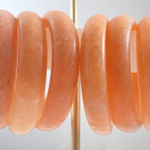 May include: Six peach-colored bangles are displayed on a stand. The bracelets have a smooth, rounded design and a translucent quality. The color is a soft, warm peach, with variations in tone. The bangles are likely made of a stone or resin material.