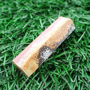 May include: A rectangular bar of soap with a pink, brown, and black marbled design. The soap is sitting on a bed of green artificial grass.