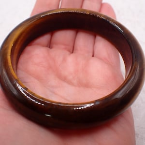 May include: A polished, round tiger's eye gemstone bangle bracelet. The bracelet is a rich brown color with golden-yellow streaks, and is approximately 2.5 inches in diameter. The smooth, glossy surface reflects light.