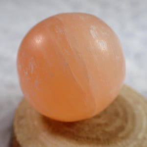 May include: A spherical, peach-coloured stone, possibly a crystal or mineral specimen. The orb rests on a small, round, wooden slice, highlighting the natural textures of both materials. The stone's surface has subtle colour variations.