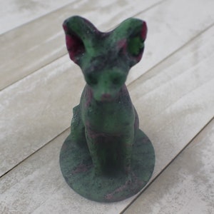 May include: Green and pink ceramic figurine of a cat sitting on a base. The cat has large ears and a textured surface.