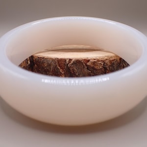 May include: A white, oval bowl with a smooth, glossy surface. Inside the bowl is a section of a tree trunk, displaying the natural wood grain and bark texture. The bowl is a decorative item.