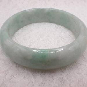 May include: A jade bangle bracelet. The bracelet is a pale green colour with white marbling throughout. The jade is polished and has a smooth, glossy finish. The bangle is a classic piece of jewellery.