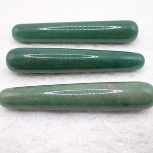 May include: Three smooth, cylindrical green jade massage tools are displayed. The jade pieces have rounded ends and a polished surface, reflecting light. The tools are arranged horizontally against a white, textured background.