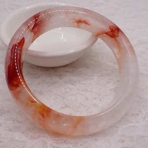 May include: A clear, circular bangle with red and orange streaks. The bracelet is displayed on a white surface. The polished bracelet is made of a smooth material. This is a piece of jewellery.