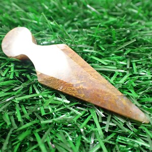 Peut inclure: Une pointe de cristal d'agate brune et blanche avec un sommet arrondi. La pointe est posée sur un lit d'herbe artificielle verte.