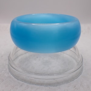 May include: A light blue, opaque, round bangle bracelet. The bracelet is a solid colour and has a smooth, polished surface. The bangle is resting on a clear glass stand.