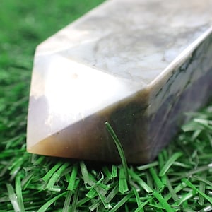 May include: A large, gray and white agate crystal point with a pointed tip. The crystal is resting on a bed of green artificial grass.