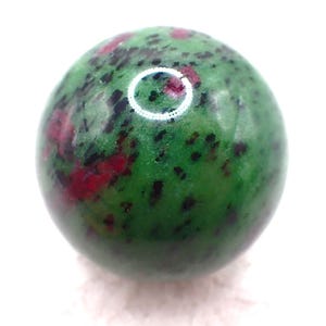 May include: A polished, spherical gemstone with a vibrant green base color. The stone is speckled with dark red and black inclusions, creating a unique pattern. The gemstone is set against a bright white background.