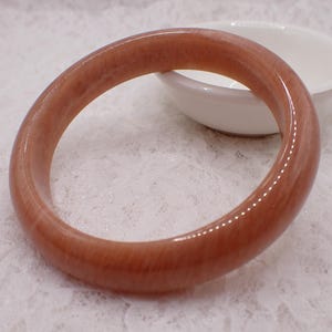 May include: A polished, reddish-brown bangle bracelet. The smooth, circular bracelet is set against a white background with a small, white, ceramic bowl. The bangle's color is a warm, earthy tone, and it appears to be made of a solid material.