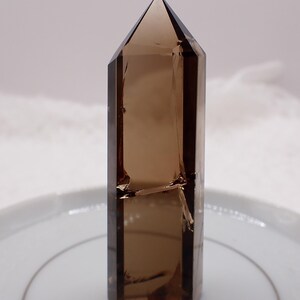 May include: A faceted, brown crystal point with a sharp, angled top. The crystal is a translucent, smoky brown color and stands upright on a white plate. The crystal has a geometric shape and appears to be a natural mineral specimen.