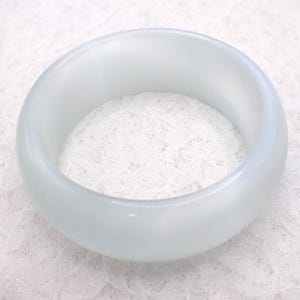 May include: A wide, light blue bangle bracelet. The bracelet is made of a smooth, opaque material and has a rounded shape. The background is a white, lacy fabric. This bracelet is a piece of jewelry.