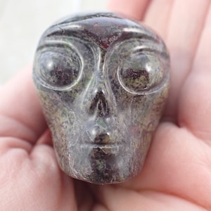 May include: A carved alien head sculpture in shades of green and brown. The sculpture has large, oval eyes, a small nose, and a defined chin. The surface has a polished, reflective quality, and the overall design is detailed and smooth.