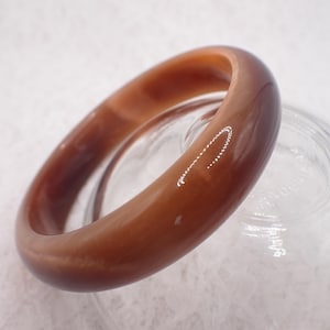 May include: A brown bangle bracelet with a smooth, polished surface. The bracelet is oval-shaped and appears to be made of a solid material. The colour varies from light to dark brown. The bracelet is resting on a clear glass surface.