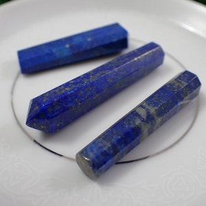 May include: Three blue lapis lazuli crystal wands on a white plate. The wands are smooth and polished with a slight sparkle.