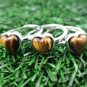 May include: Three adjustable silver rings, each featuring a heart-shaped tiger's eye stone. The rings are displayed on a bed of green artificial grass, creating a natural contrast with the polished metal and warm-toned gemstones.
