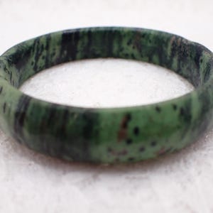 May include: A green and black speckled jade bangle bracelet. The bracelet is a solid band of polished stone, showcasing a natural pattern of dark green and black hues. The bracelet is approximately 6.35 cm in diameter.