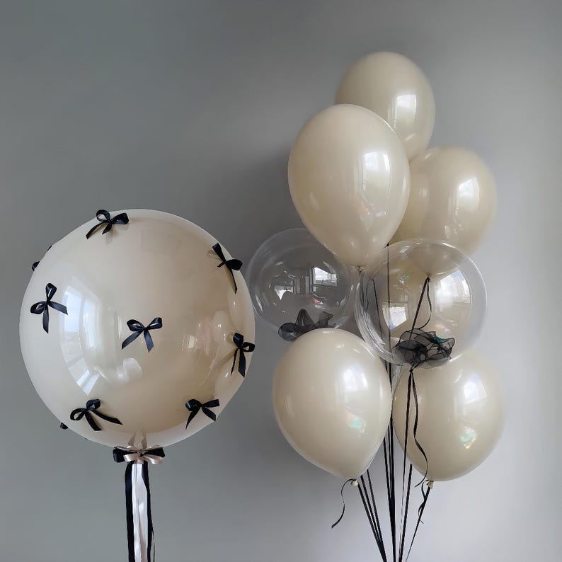May include: A group of balloons in various sizes and colours. The balloons are tied together with black ribbons. Some of the balloons are clear and have black bows inside. The balloons are on a white background.