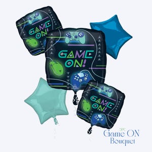 May include: A collection of five foil balloons in various shapes. The central balloon is square and features the words "GAME ON!" along with images of a game controller and mouse. Other balloons are star-shaped and diamond-shaped, in shades of blue and teal.