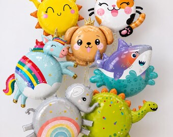 May include: A collection of round foil balloons featuring cartoon characters including a yellow sun, a calico cat, a tan puppy with a crown, a blue unicorn with a rainbow, a purple shark, a gray elephant with a rainbow, and a green dinosaur.