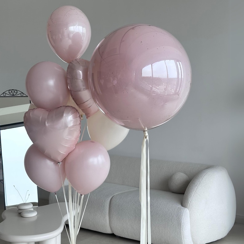May include: A collection of pink balloons, including round and heart-shaped designs, tied together with white ribbons. The balloons vary in size and shade, creating a festive and celebratory display. A large, clear balloon with pink confetti is a focal point.