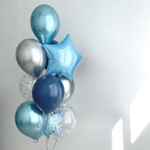 May include: A balloon bouquet featuring a mix of metallic light blue, dark navy blue, and silver round balloons, a single light blue star-shaped foil balloon, and two clear balloons filled with silver confetti, all tied together with thin white ribbons.