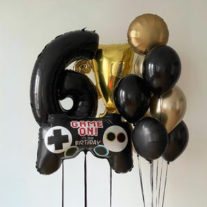 May include: A black and gold balloon bouquet with a large black balloon shaped like a video game controller. The controller has the text "Game On! It's your birthday" printed on it. The bouquet also includes a gold trophy balloon and several black and gold balloons.