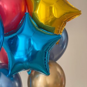 Puede incluir: Un grupo de globos coloridos, que incluyen un globo rojo, un globo azul en forma de estrella, un globo dorado en forma de estrella y un globo dorado.
