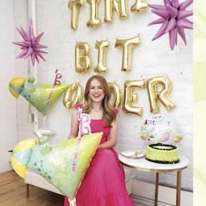 May include: A woman in a pink dress smiles, holding a cocktail-shaped balloon. Gold letter balloons spell "TINY BIT OLDER" on the white brick wall. A small cake and champagne glasses sit on a table. Decorative starburst balloons are also present.