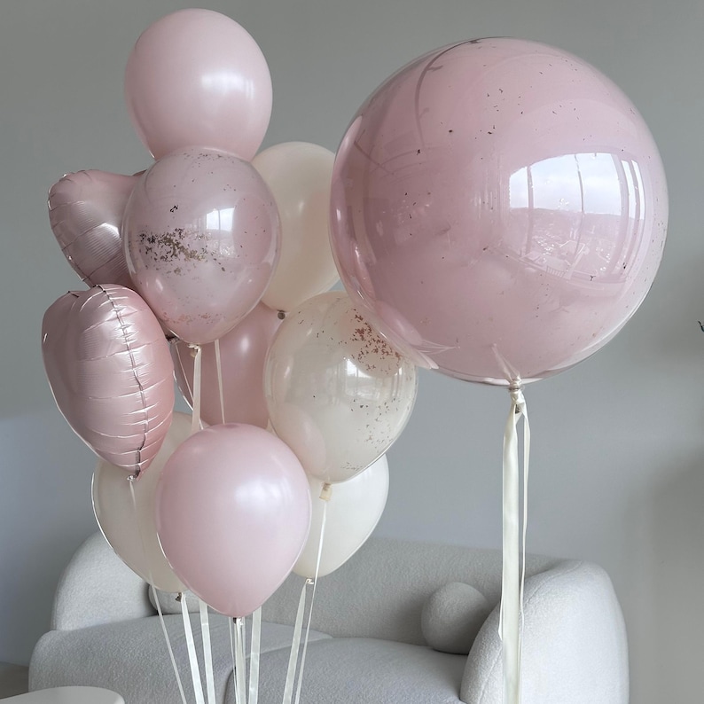 May include: A collection of pink and white balloons, some with confetti, tied together with white ribbons. The balloons vary in size and shape, including round and heart-shaped. The largest balloon is a clear bubble with pink confetti.