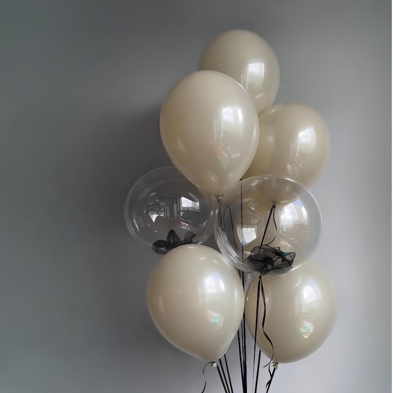 May include: A bunch of cream-coloured balloons with black ribbon ties. Some of the balloons are clear and have black tulle inside.