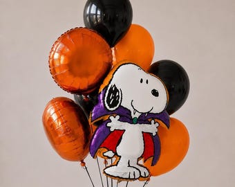 May include: A balloon bouquet featuring a large foil balloon shaped like Snoopy dressed as a vampire with a purple and red cape, surrounded by round orange and black latex and foil balloons. The arrangement is tied together with thin black strings.