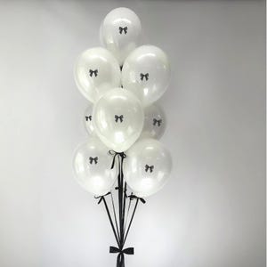 May include: A cluster of eight round, white balloons, each featuring a small, centered black bow graphic. The balloons are tied together at the base with long, thin black ribbons that extend downward, creating a cohesive decorative arrangement.