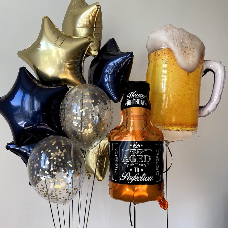 Op de afbeelding: Een bos ballonnen voor een verjaardagsfeest. De ballonnen zijn goud, zwart en transparant met gouden confetti. De ballonnen zijn in de vorm van sterren, een bierpul en een fles drank. De fles drank heeft de tekst "Happy Birthday" en "Aged to Perfection".