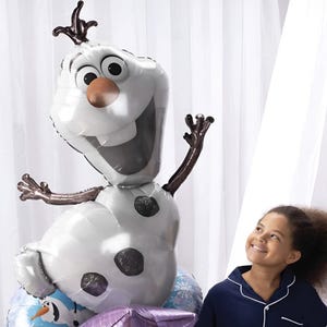 May include: A large, white Olaf balloon with a brown nose, black eyes, and a wide smile. The balloon has dark brown twig-like arms and a crown. A young girl in blue pajamas looks up at the balloon with a smile.