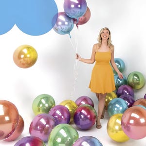 May include: A woman in a yellow dress holds a bouquet of iridescent balloons. The balloons are scattered on the floor and floating in the air. The balloons have a metallic finish and reflect colors like purple, green, and gold.
