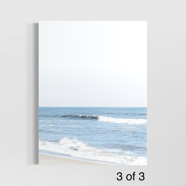 May include: A framed print featuring a serene beach scene. The image captures the ocean with gentle waves, a sandy shore, and a clear sky. The color palette is dominated by shades of blue and white, creating a calming aesthetic. The text "3 of 3" is visible at the bottom.