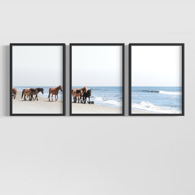 May include: Set of three framed art prints featuring a beach scene. The prints depict horses of varying brown and black hues walking along a sandy beach with the ocean in the background. The sky is a pale blue.