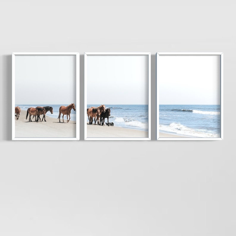 May include: Triptych of framed art prints featuring wild horses on a sandy beach. The horses, in shades of brown and black, are near the water's edge. The ocean and sky are a soft blue, creating a serene coastal scene.